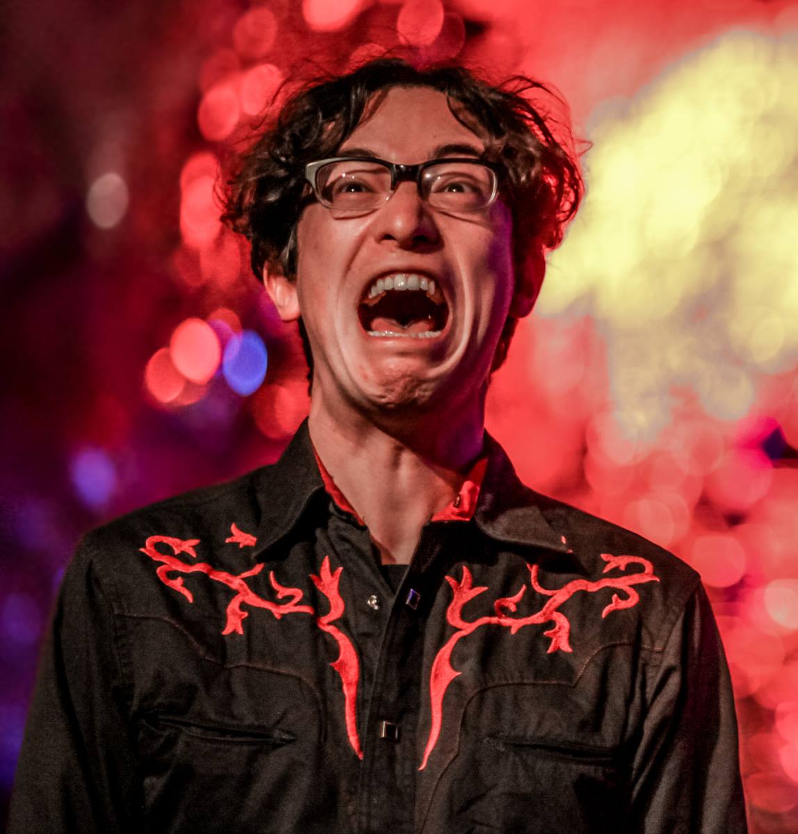 Jeffrey Young screaming during a performance, wearing an elaborate western shirt in front of a red background