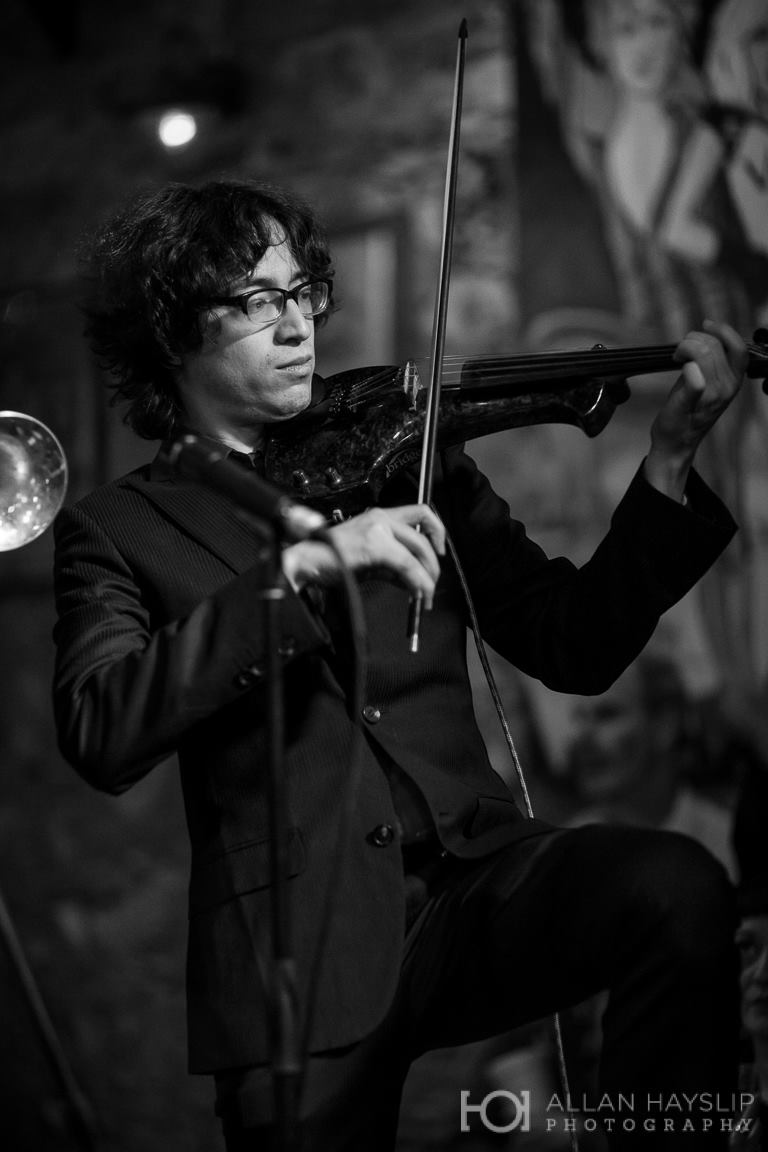 Black and white photo of Jeffrey Young performing on a solid-body electric violin with one knee raised while wearing a suit