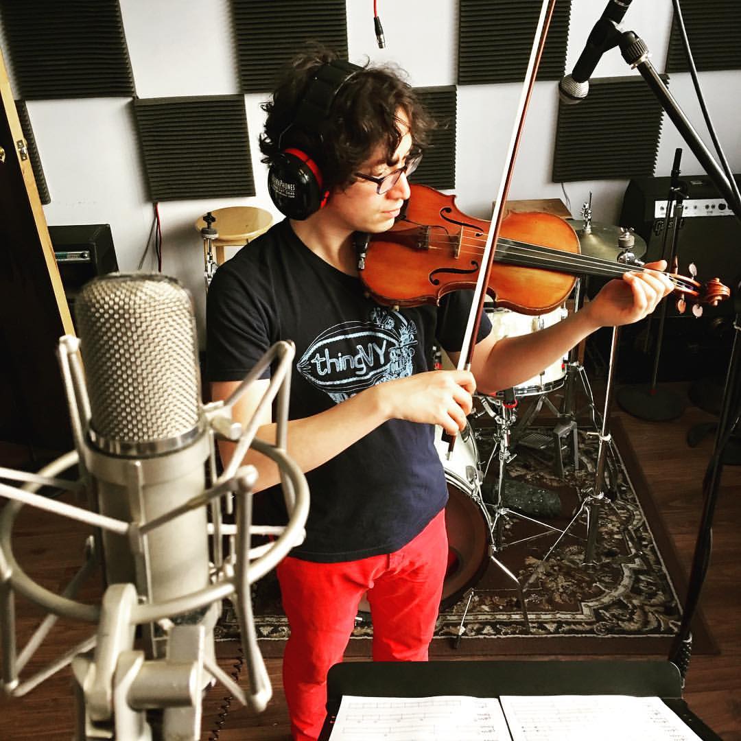 Jeffrey Young playing violin in a recording studio, standing in front of a silver condenser mic with a drumset and a black and white wall in the background