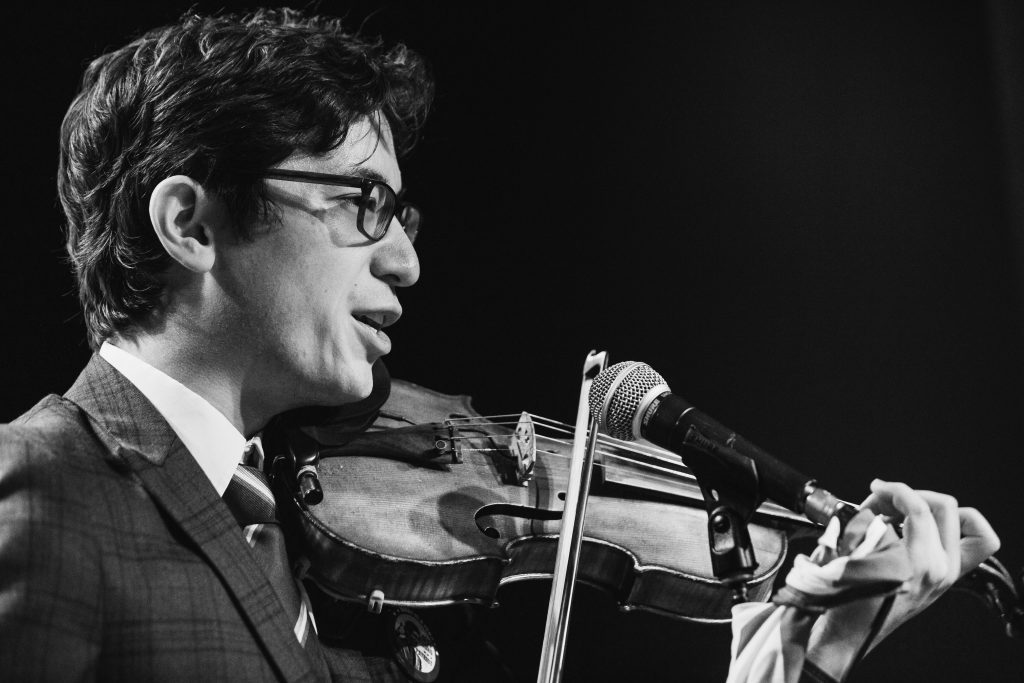 Black and white photo of Jeffrey Young performing the Patriots experimental opera, speaking into a mic while playing violin