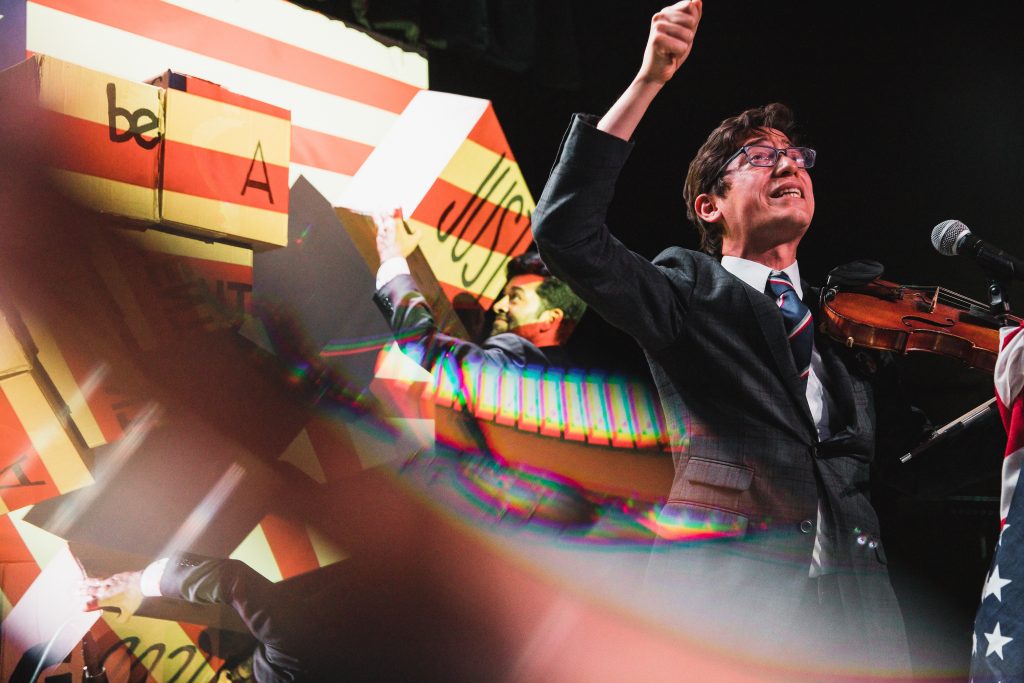 Jeffrey Young performing the Patriots experimental opera, with Paul Pinto in the background rearranging boxes with words on them and a distorted lens effect on the photo