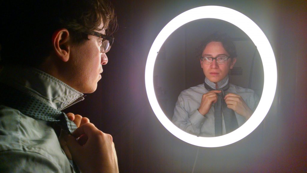 Jeffrey Young stares moodily at his reflection in a circular mirror rimmed with bright lights while tying a tie