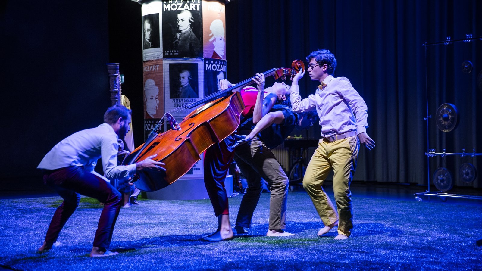 Jeffrey Young and others performing NOMOZART in Switzerland, forming an elaborate group pose while levitating an upright bass, in front of a set including a European-style advertising pole with images of Mozart