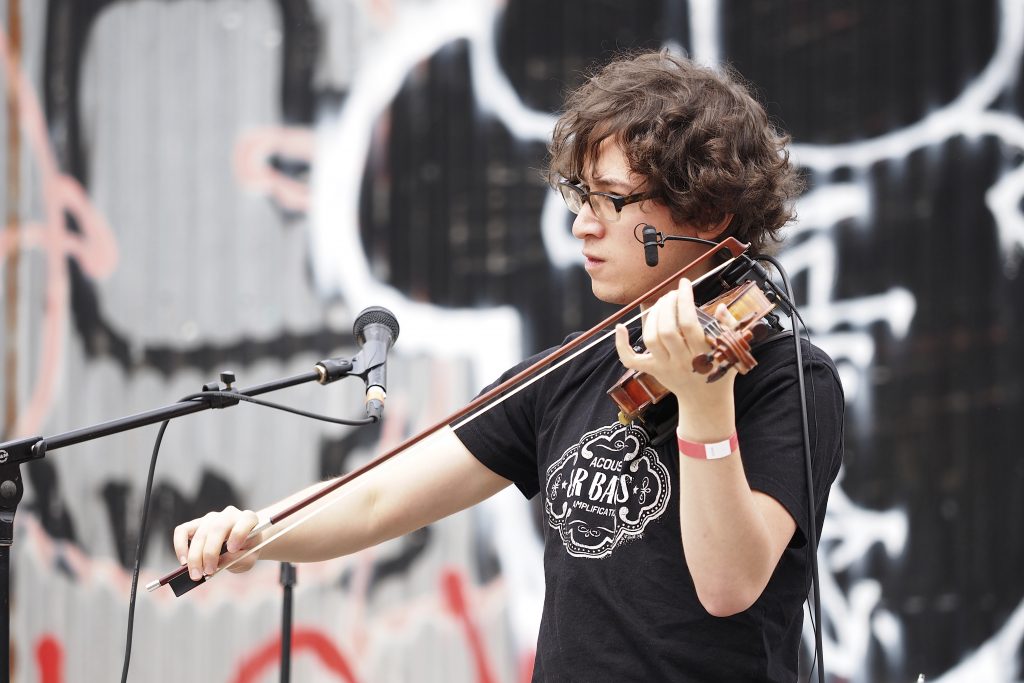 Jeffrey Young performing outdoors in a T-shirt at Bushwick Open Studios in front of a graffiti-tagged wall