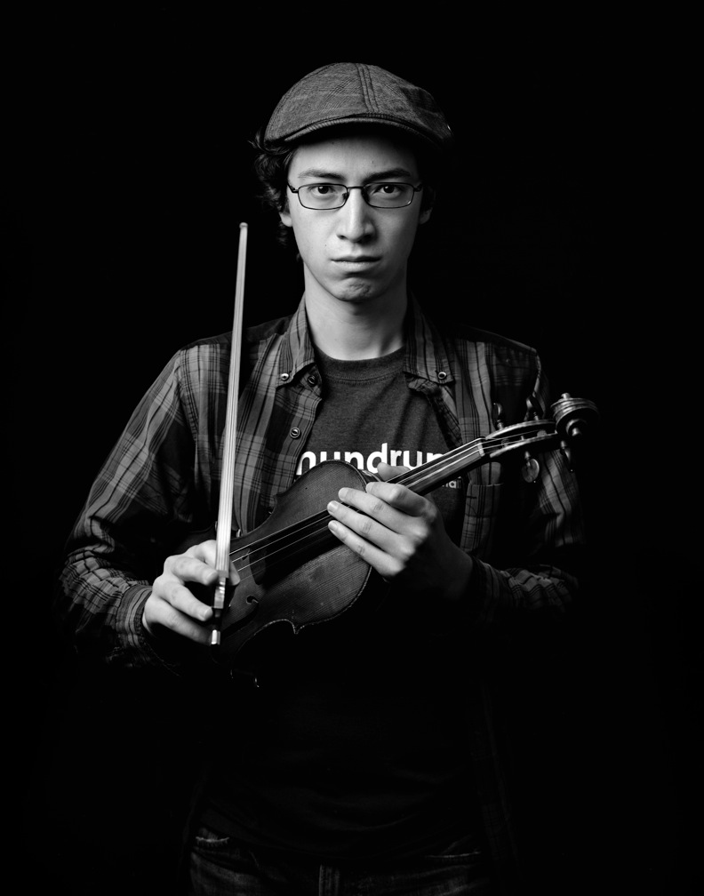 Black and white headshot of a younger Jeffrey Young in a newsboy cap with violin tucked under an arm and bow slung over the shoulder, with an intense expression