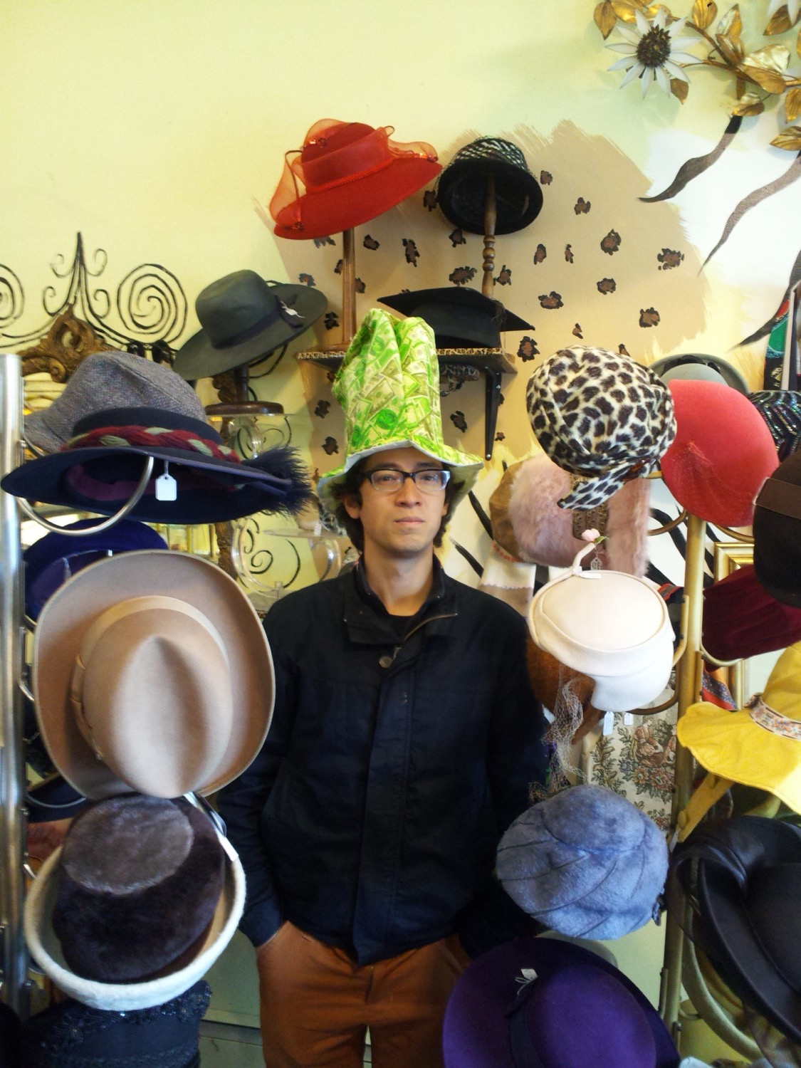 Jeffrey Young with a bemused expression wearing a tall hat with dollar bills pictured on it, surrounded by hats of all kinds