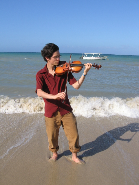 Jeffrey Young playing violin with windswept hair while standing in the tide, with the ocean and a boat in the background