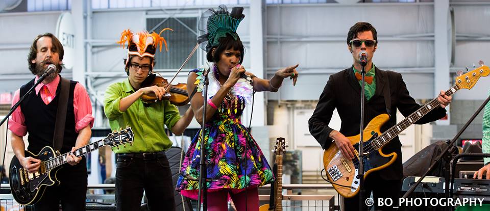 Jeffrey Young performing while wearing an elaborate headdress with a band wearing elaborately colored clothing