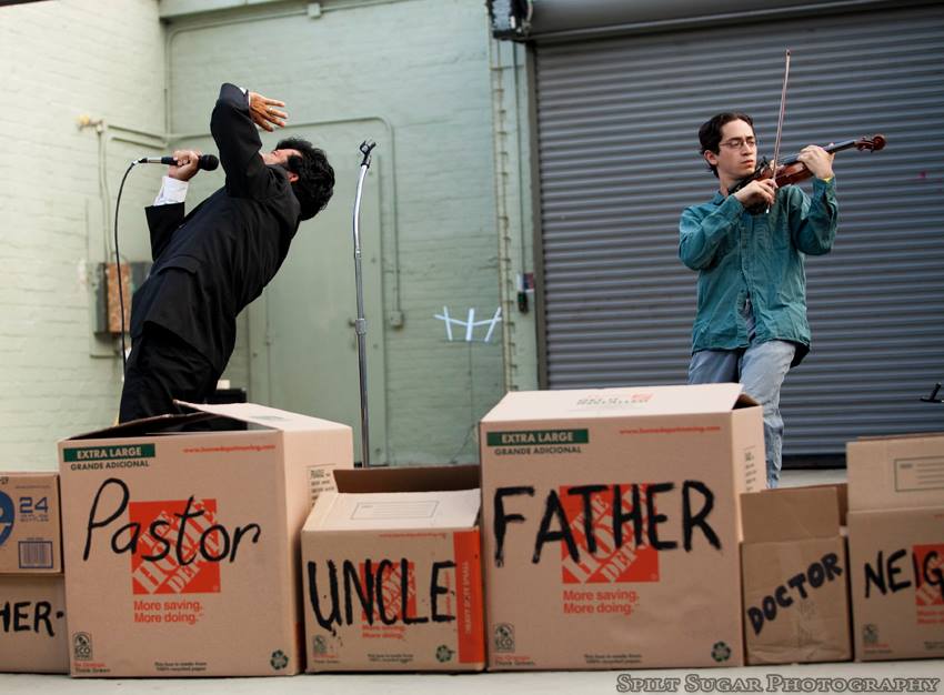 Jeffrey Young performing the Patriots experimental opera on violin while Paul Pinto shouts into a mic in the foreground, all behind a wall of cardboard boxes with words on them including Pastor, Uncle, and Father