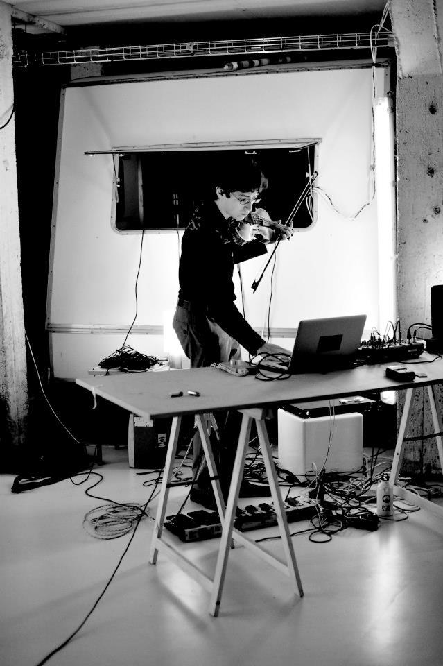 Black and white photo of Jeffrey Young performing on violin and laptop in an underground venue in Paris, France, with a trailer behind him in a large concrete space