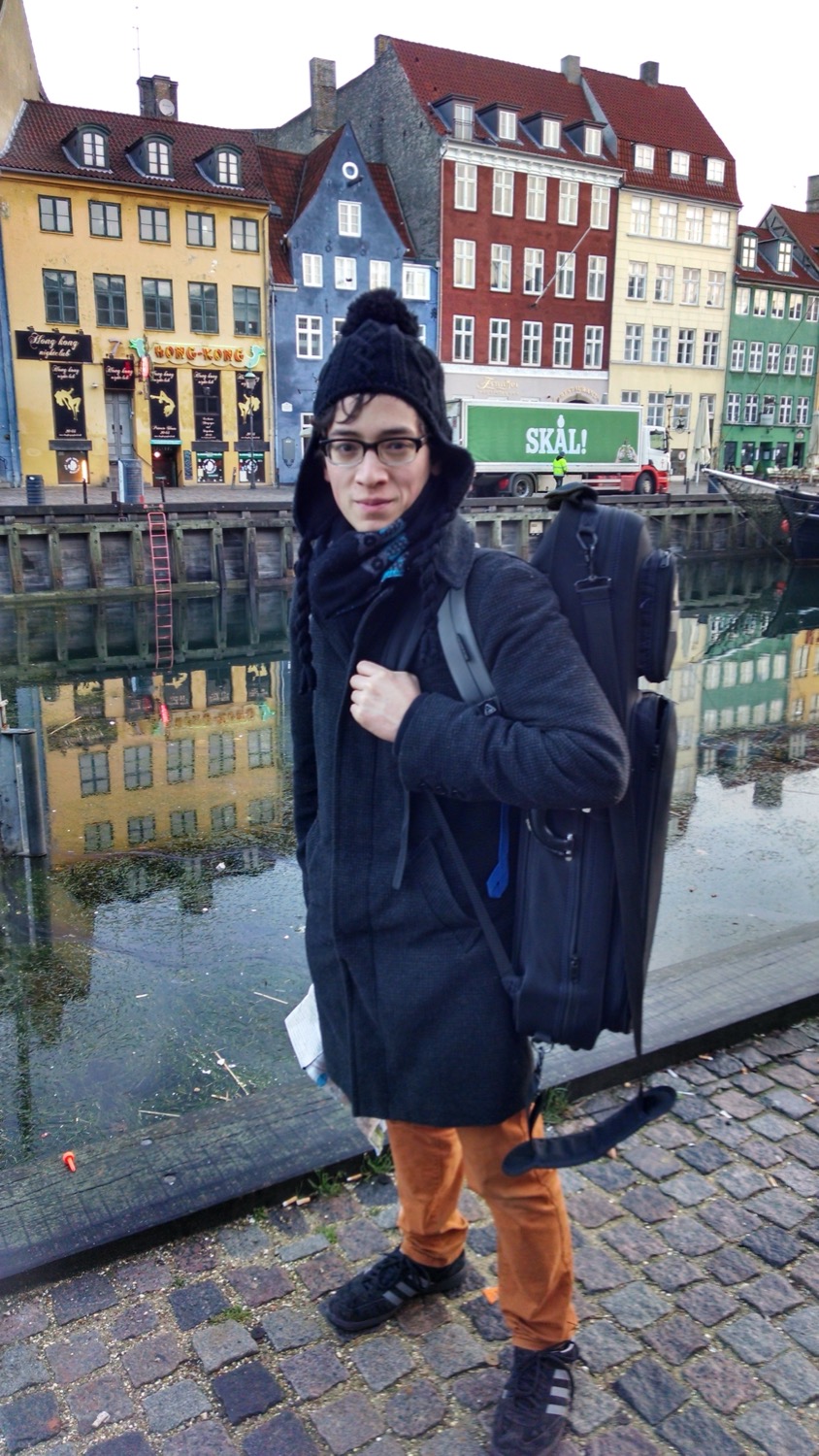 Jeffrey Young in winter clothes including a hat with a pom-pom, wearing his violin in its case on his back in front of a canal in Copenhagen, Denmark, featuring a truck in the background on which is written Skål!