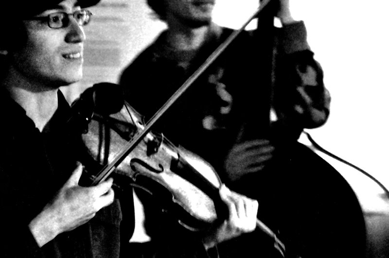 Black and white photo of Jeffrey Young looking up with a smile while holding his violin, with a bassist in the background
