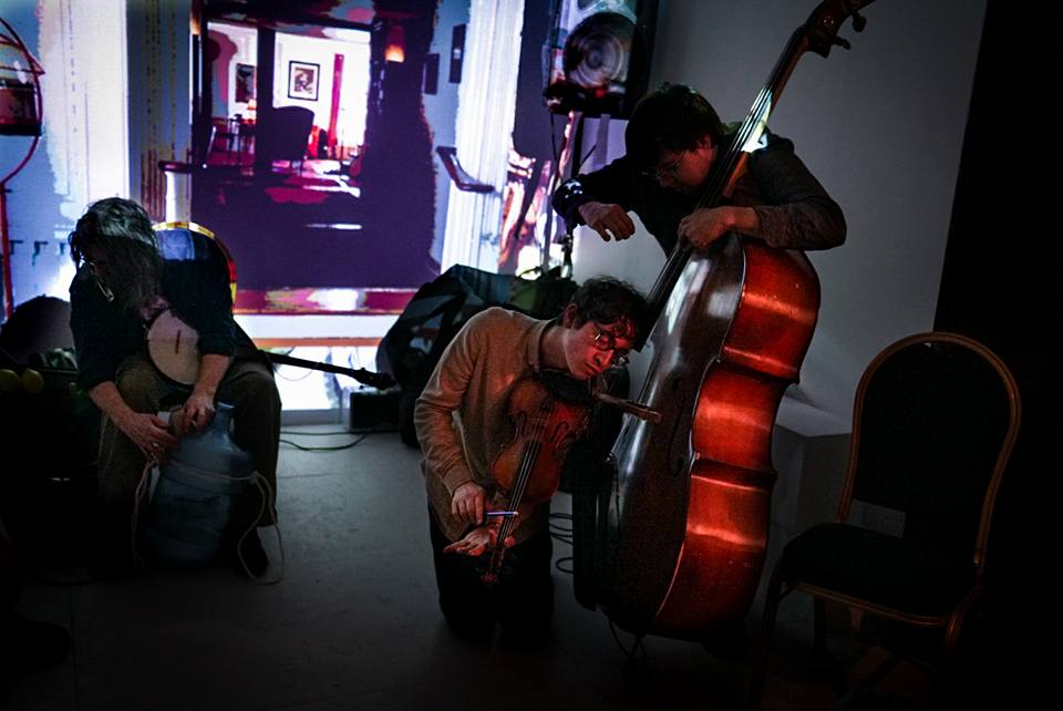 Jeffrey Young performing at XFest in New England, on his knees with violin pointed down and eyes closed, leaning sensuously against the strings of an upright bass as another person plays it, with a projection in the background