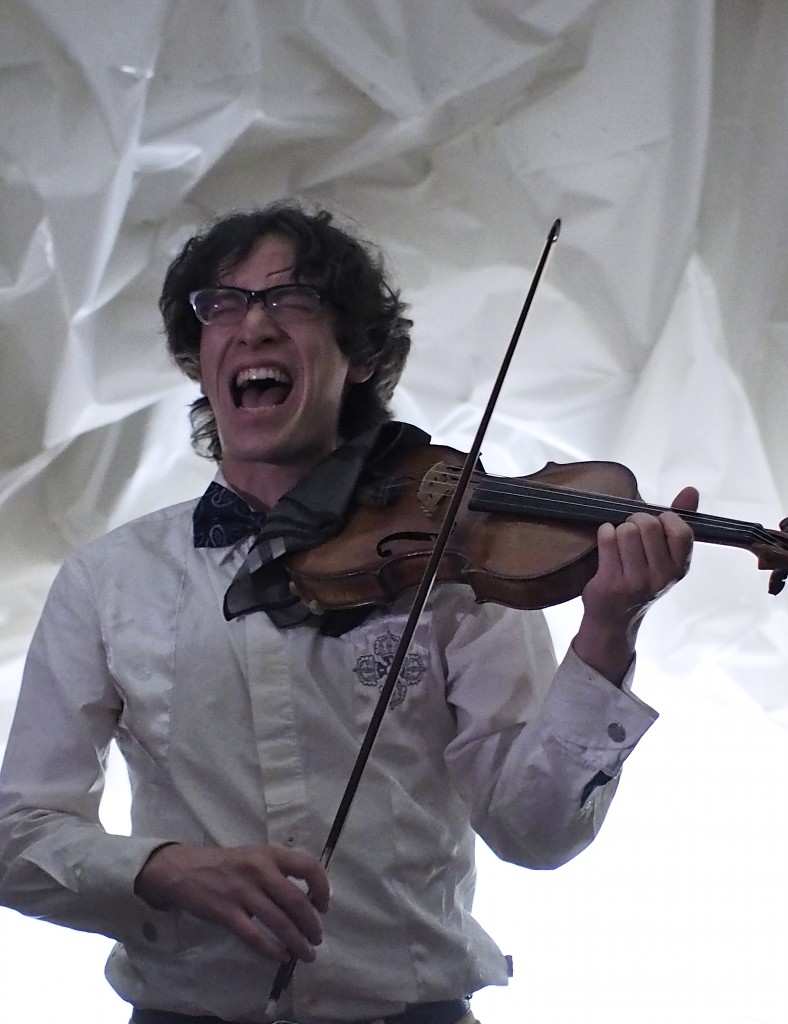 Jeffrey Young performing on violin while screaming, wearing a silken white shirt and bowtie in front of a white crinkled fabric background